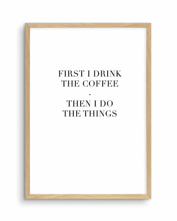 Framed art print with natural wood border displaying black serif text on white background. Text reads "FIRST I DRINK THE COFFEE - THEN I DO THE THINGS" arranged in three centered lines. Clean, minimalist design with classic typography and simple composition.