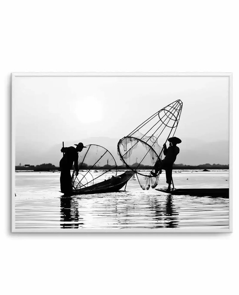 Framed black and white photograph of two fishermen in a lake with large, conical fishing nets, displayed in a white frame with a white border. The composition features the silhouettes of two men, one standing in a small boat and the other standing in the water, both holding their nets. The background shows a hazy horizon with distant mountains under a bright sky, and the foreground captures the rippling water.