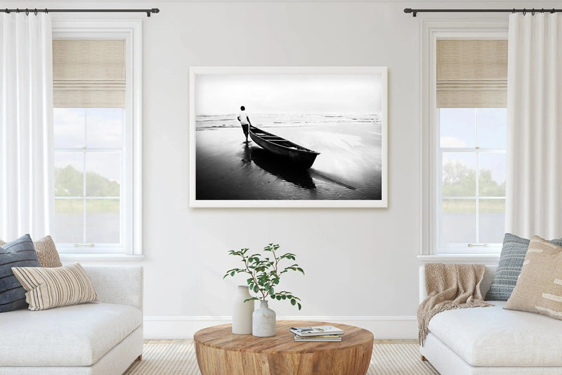 Lifestyle shot of a minimalist living room featuring a large framed black and white photograph of a coastal beach scene displayed above a round wooden coffee table. The print features a person pushing a small boat into the water at low tide, with gentle waves and an overcast sky stretching to the horizon, framed in a white frame with a white border. The room includes two white sofas with textured pillows and a cream rug.