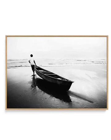 Black and white photograph in natural wood frame showing a shirtless fisherman standing beside a dark wooden boat on a wet beach. The man holds the boat's rope while facing the calm ocean horizon. Wet sand reflects the figure and vessel. Overcast sky creates a muted, contemplative atmosphere. Fine art photography print with subtle gradient tones from dark blacks to light grays. Natural wood frame with white border.