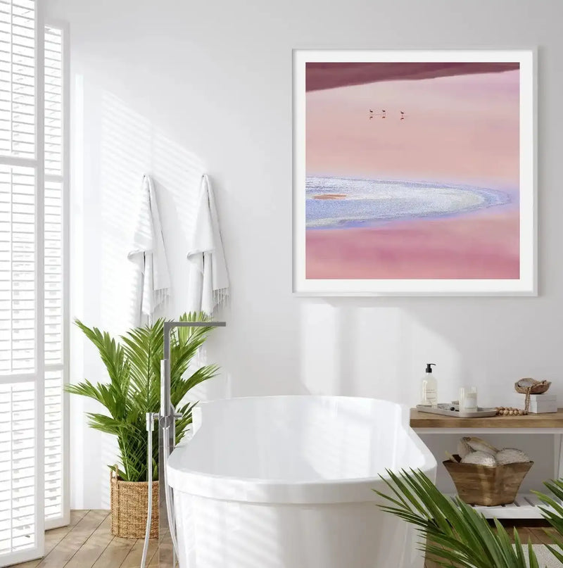 Lifestyle shot of a modern bathroom featuring a large framed art print above a white freestanding bathtub. The print depicts an aerial view of a pink salt lake with four small flamingo figures in the upper left, with a white frame and white border. The bathroom has white walls, a large window with white blinds, a potted palm plant in a woven basket, and a wooden shelf with bath accessories.