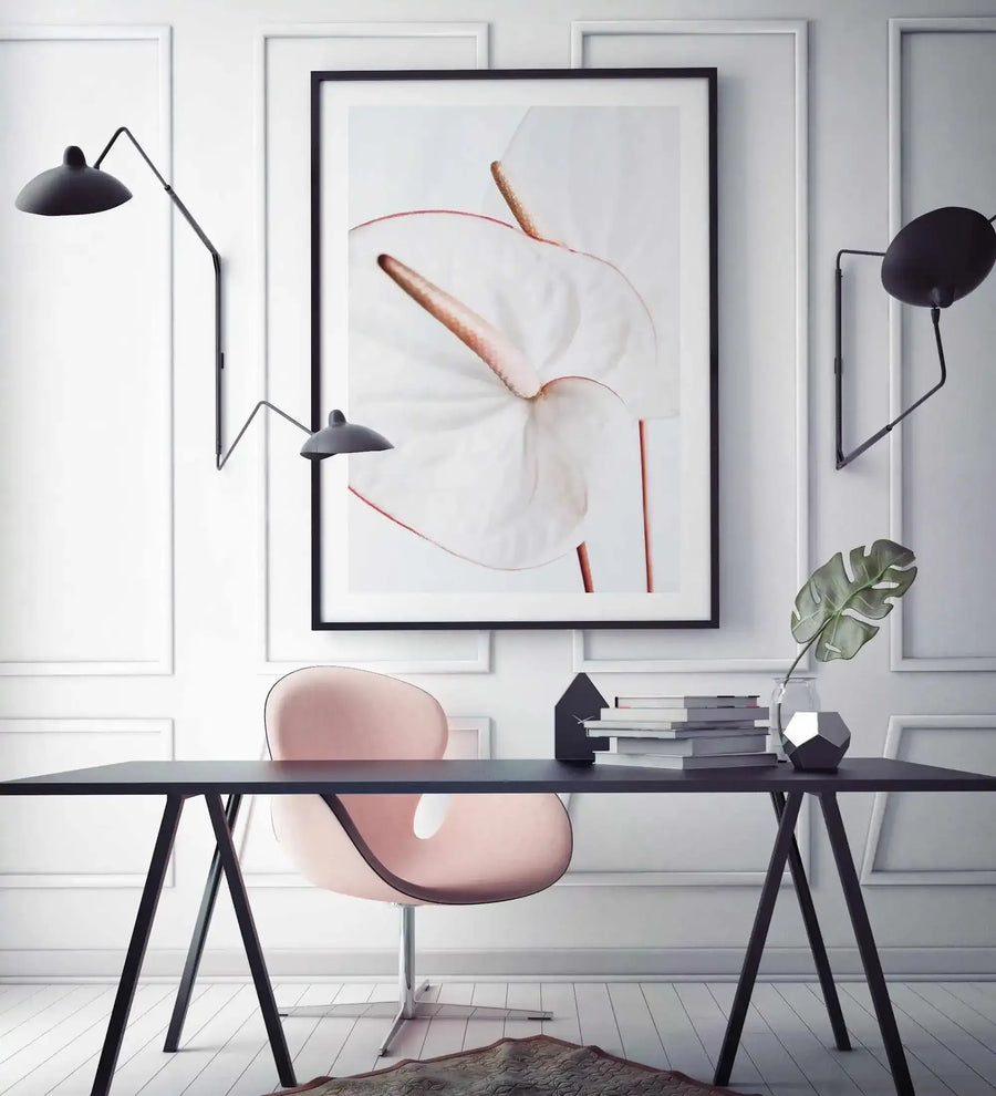 Framed art print titled Fleur Flamingo displayed above a black desk in a minimalist office setting. The artwork features an abstract watercolor illustration with soft blush pink and terracotta tones, depicting stylized botanical forms and curved lines suggesting a flamingo silhouette. The piece is mounted in a black frame with white border against white paneled walls. A pink upholstered chair, black arc lamp, and potted monstera plant complete the workspace styling.
