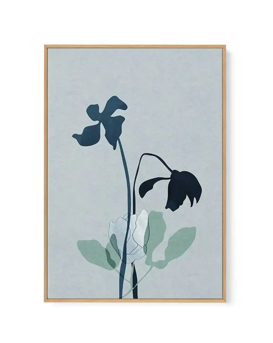 Framed canvas art print featuring a minimalist botanical illustration against a soft pale blue background. Two delicate flowers with dark teal-blue petals are rendered in silhouette, one upright and one drooping downward. Thin dark stems connect to layered green and white leaves below in muted sage and pale green tones. The composition is vertically oriented and centered. The canvas is set in a natural wood frame with a subtle shadow effect visible on the right edge.
