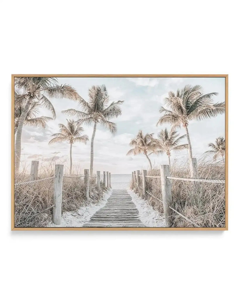 Framed canvas art print depicting a wooden boardwalk pathway leading toward calm ocean waters. Tall palm trees with light tan and green fronds frame both sides of the composition, with dried beach grasses and wooden fence posts lining the walkway. The sky features soft white and gray clouds. The artwork has a serene, coastal aesthetic rendered in muted, naturalistic tones of cream, beige, gray, and soft green. The canvas is mounted in a natural wood frame with a white border.