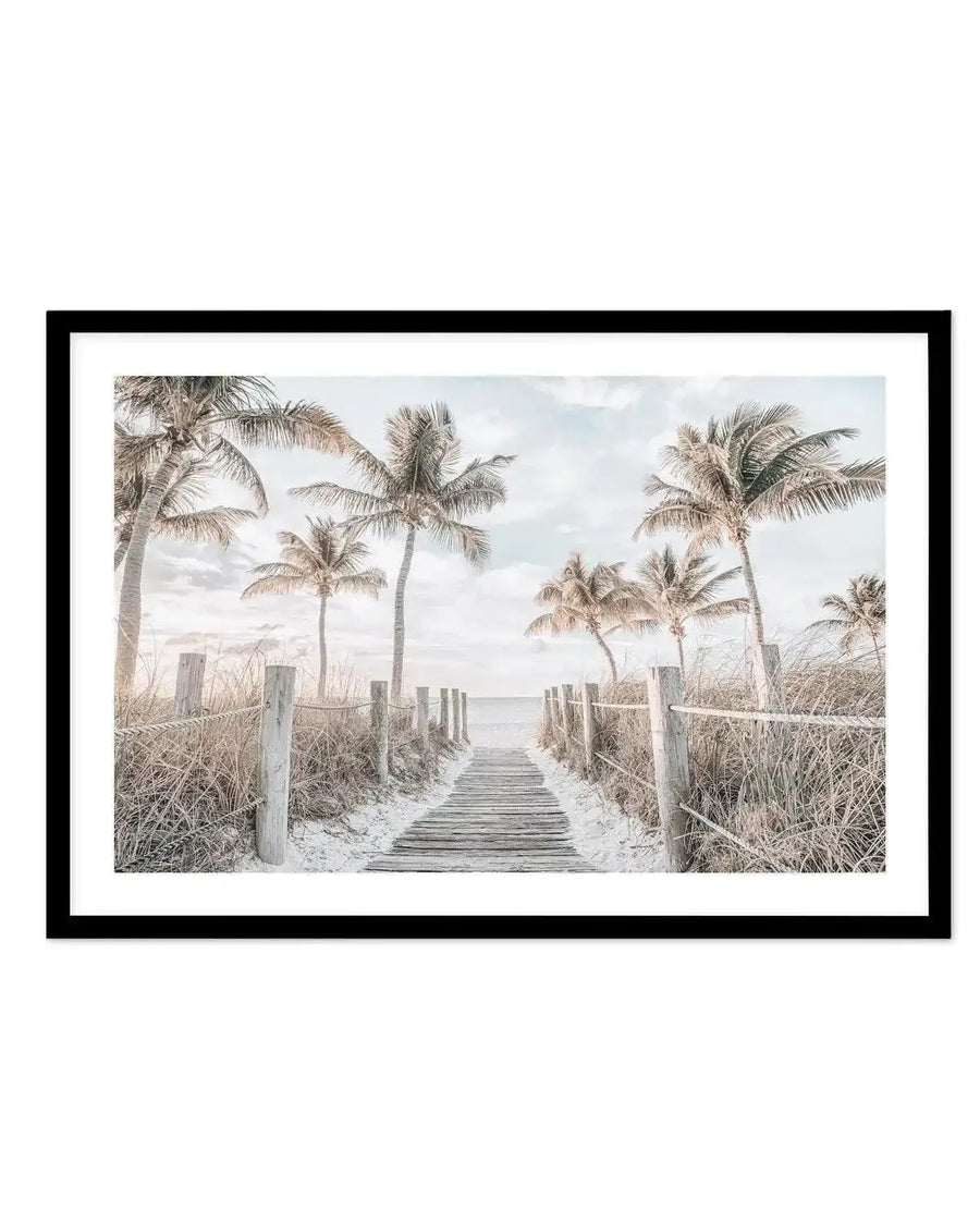Framed photography art print of a serene beach scene with a wooden boardwalk leading to the ocean, displayed in a black frame with a white border. The composition features numerous palm trees with light green and muted brown fronds under a soft blue and white cloudy sky, with dry, light brown grasses lining the boardwalk.