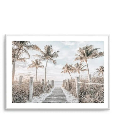 Framed photography art print of a serene beach scene with a wooden boardwalk leading to the ocean, framed in white with a white border. The composition features tall palm trees with light green fronds on either side of the boardwalk, with dry, light brown beach grass and wooden posts with rope fencing. The sky is a soft blue with scattered white clouds, and the overall mood is calm and inviting.