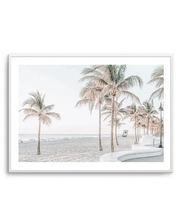 Framed photography art print of a serene beach scene with a row of palm trees and white beach chairs under a pale sky, with a white frame and white border. The composition features a sandy beach with gentle waves in the distance, a white lifeguard stand, and a curved white concrete wall with a lamppost on the right.