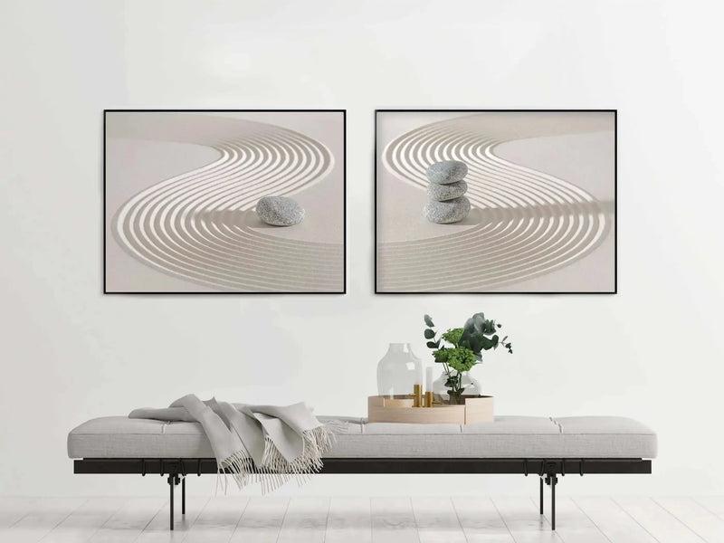 Lifestyle shot of two framed art prints displayed side-by-side above a long, light grey upholstered bench in a minimalist room. The prints feature a zen garden with raked sand patterns and stacked stones in muted grey and cream tones, with black frames and no border. A light grey throw blanket with fringe is draped over the left side of the bench, and a wooden tray with two clear glass vases, two gold candle holders, and a vase with green foliage sits on the right.