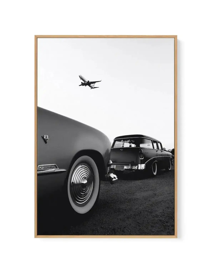 Framed black and white photograph of two vintage cars and an airplane in a natural wood frame with white border. The composition features the front quarter of a classic car with a whitewall tire and chrome hubcap in the foreground, with another vintage station wagon parked behind it. An airplane is visible in the sky above the cars, taking off against a bright white sky.