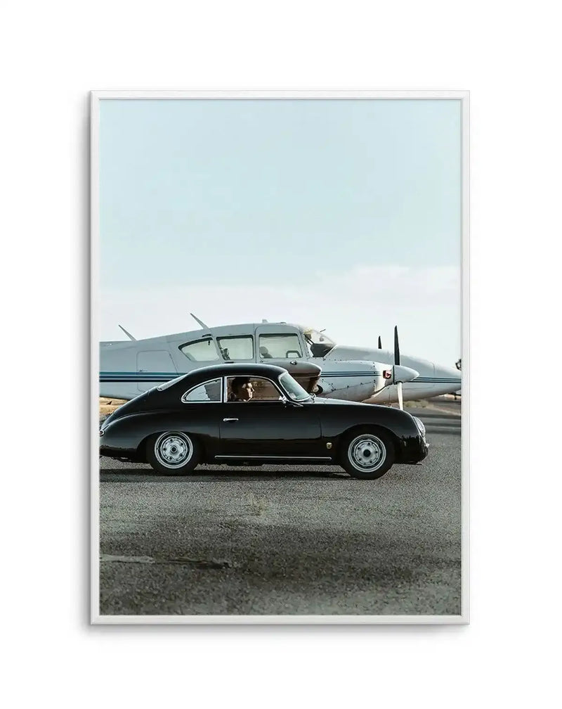 Framed black and white photograph of a vintage black Porsche 356 parked on an asphalt runway with a small white propeller plane in the background, under a pale blue sky. The car is positioned in the mid-ground, facing right, with a person visible in the driver's seat. The plane's tail and propeller are visible behind the car.