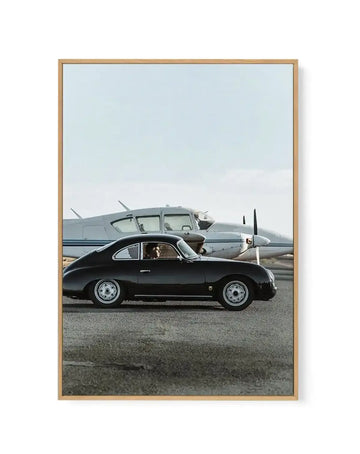 Framed canvas art print titled "Fly With Pam" by Tim Harris featuring a black vintage Porsche 356 coupe positioned in profile on an airport tarmac. A man sits in the driver's seat visible through the side window. Behind the car, a silver twin-engine propeller airplane is parked on the runway under a pale blue sky with light clouds. The photograph has a mid-century aesthetic with muted, naturalistic colors. The canvas is mounted in a natural wood frame with a white border.