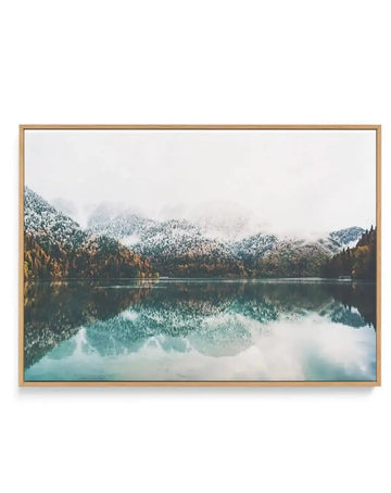 Gallery-wrapped canvas print of a serene winter landscape with a lake reflecting snow-covered mountains and an overcast sky, displayed in a natural oak frame. The mountains are covered in white snow and dark green and brown trees, with a misty, light grey sky above. The lake's surface is calm, mirroring the mountains and sky in shades of deep teal, grey, and white.