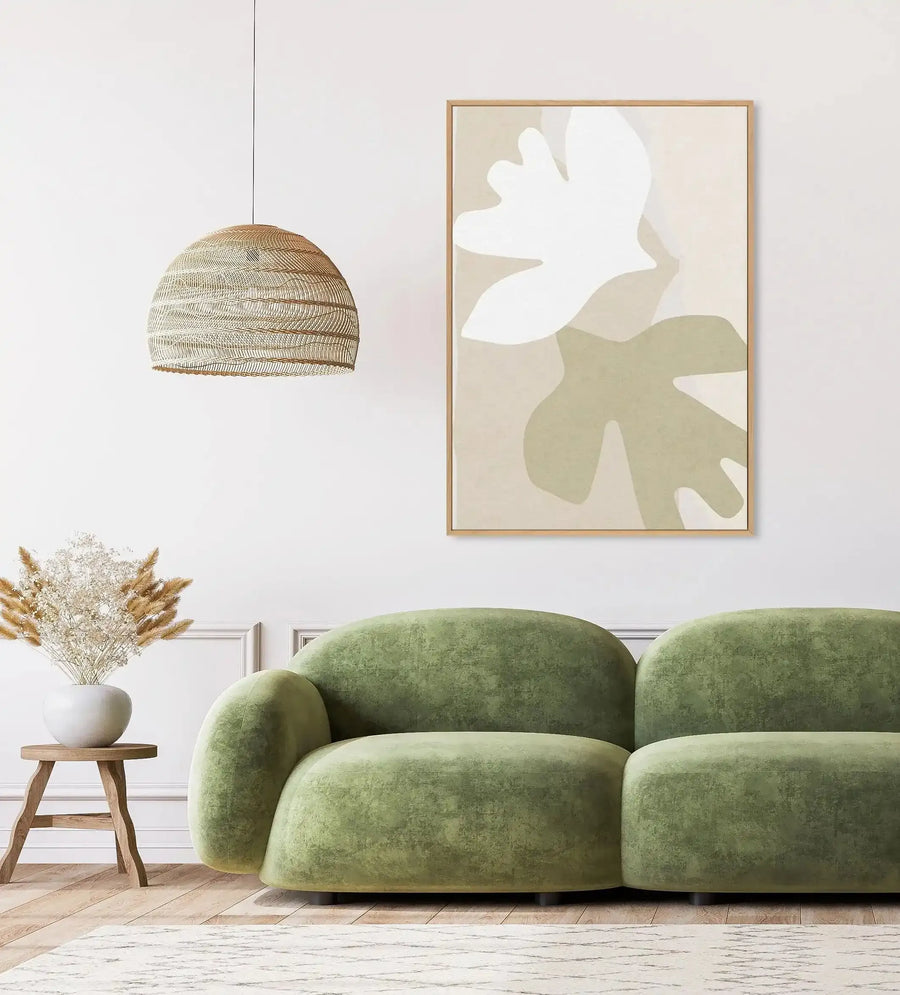 Large framed canvas art print displayed above a curved green sofa in a minimalist living room. The print features two abstract, organic shapes resembling leaves or birds, one in white and one in muted sage green, against a warm beige background, with a natural wood frame. The room includes a woven pendant light, a small wooden side table with a white vase holding dried pampas grass and baby's breath, and a textured cream rug on a light wood floor.