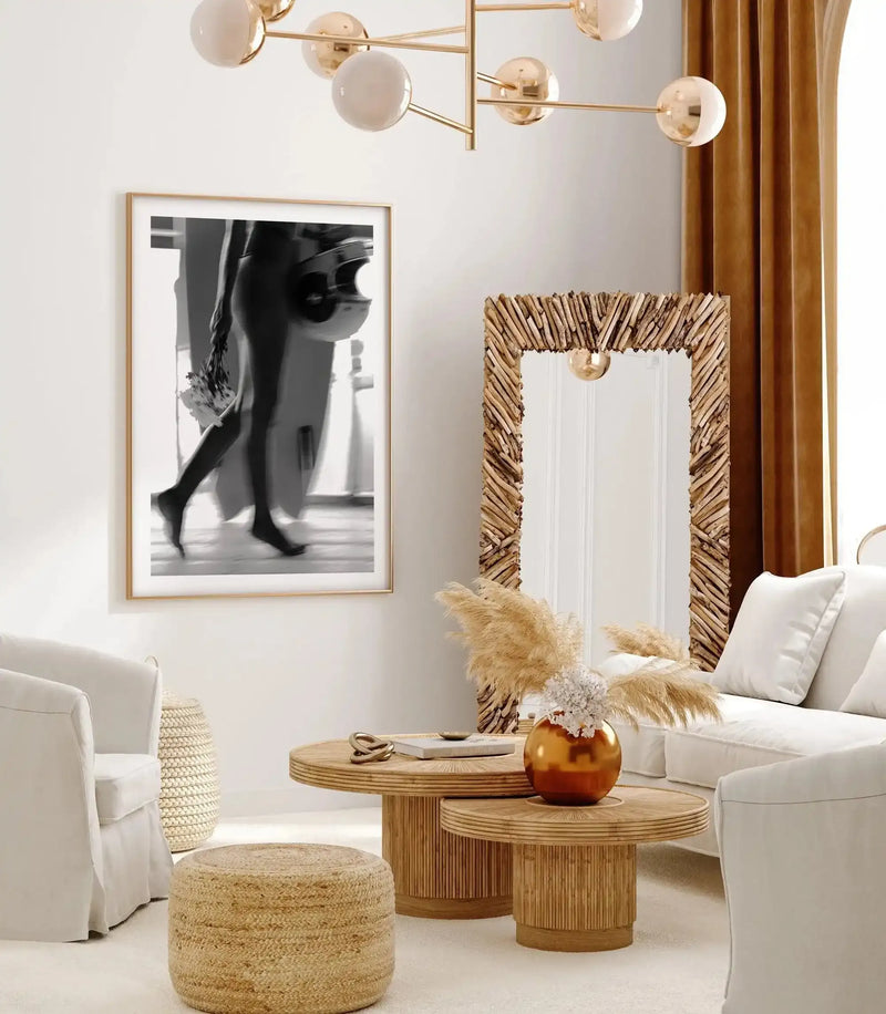 Black and white art print by Mario Stefanelli titled "Fresh as a Daisy" displayed in a natural wood frame with white border, shown in a modern living room setting alongside a woven mirror with natural wood frame, wooden side tables, cream-colored upholstered seating, a gold sphere vase with dried pampas grass, and brass globe pendant lighting.