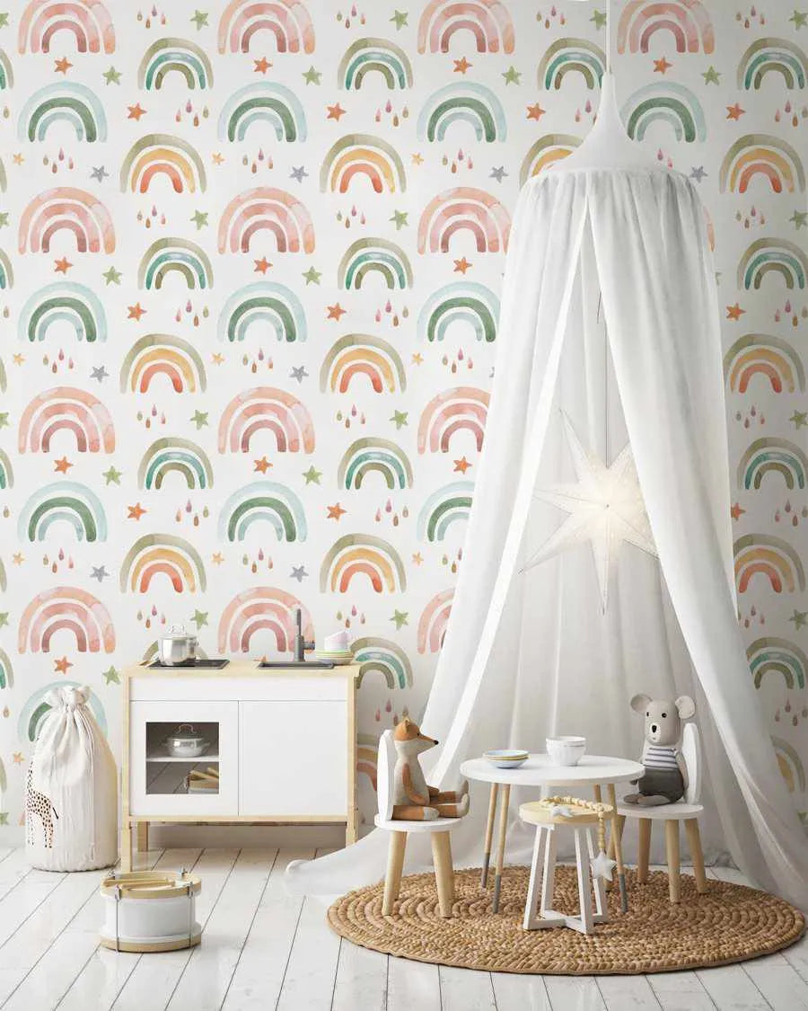 Lifestyle shot of a child's room featuring Fun Rainbows I wallpaper. The wallpaper has a repeating pattern of hand-painted watercolor rainbows in muted shades of terracotta, sage green, blush pink, and pale yellow, with small stars and raindrops scattered between them on a white background. The room includes a white play kitchen, a white canopy over a small round table with two chairs, and a woven rug.