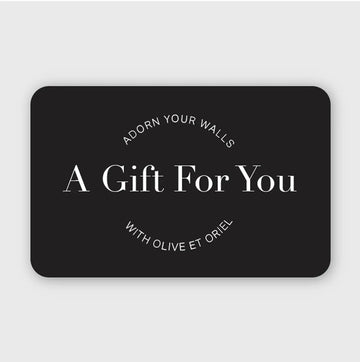 Black rounded rectangle gift card on light gray background featuring white serif and sans-serif typography. Text reads "ADORN YOUR WALLS" in curved text at top, "A Gift For You" in large centered serif font, and "WITH OLIVE ET ORIEL" in curved text at bottom. Minimalist, elegant design with high contrast black and white color scheme.