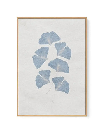Framed canvas art print featuring a botanical illustration of ginkgo leaves in soft dusty blue arranged vertically along a delicate stem on a cream background. The leaves are rendered with fine linear detailing showing their characteristic fan shape. The artwork is mounted in a natural wood frame with a white border.
