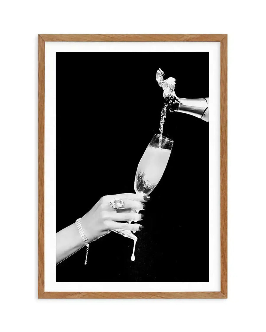 Black and white photograph featuring a woman's hand holding a champagne flute with effervescent liquid splashing upward as champagne is poured from a bottle held above. The woman wears a pearl bracelet and has manicured nails. The composition is shot against a solid black background, creating high contrast. The artwork is displayed in a natural wood frame with a white border.