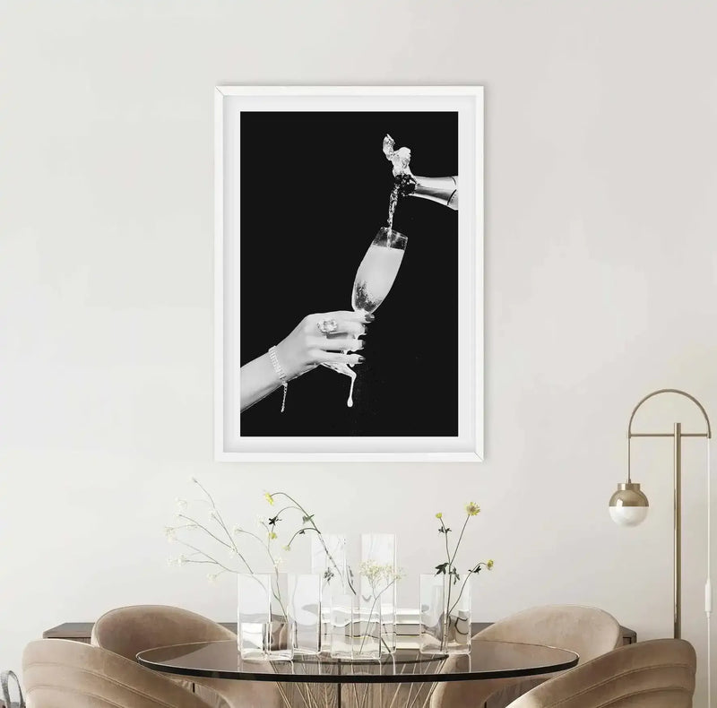 Black and white photograph showing a hand with a bracelet holding a champagne flute while champagne is being poured into it against a dark background. The image is framed in a white frame with white border, displayed above a round black table with fresh flowers in glass vases and modern tan leather chairs in a minimalist room setting.