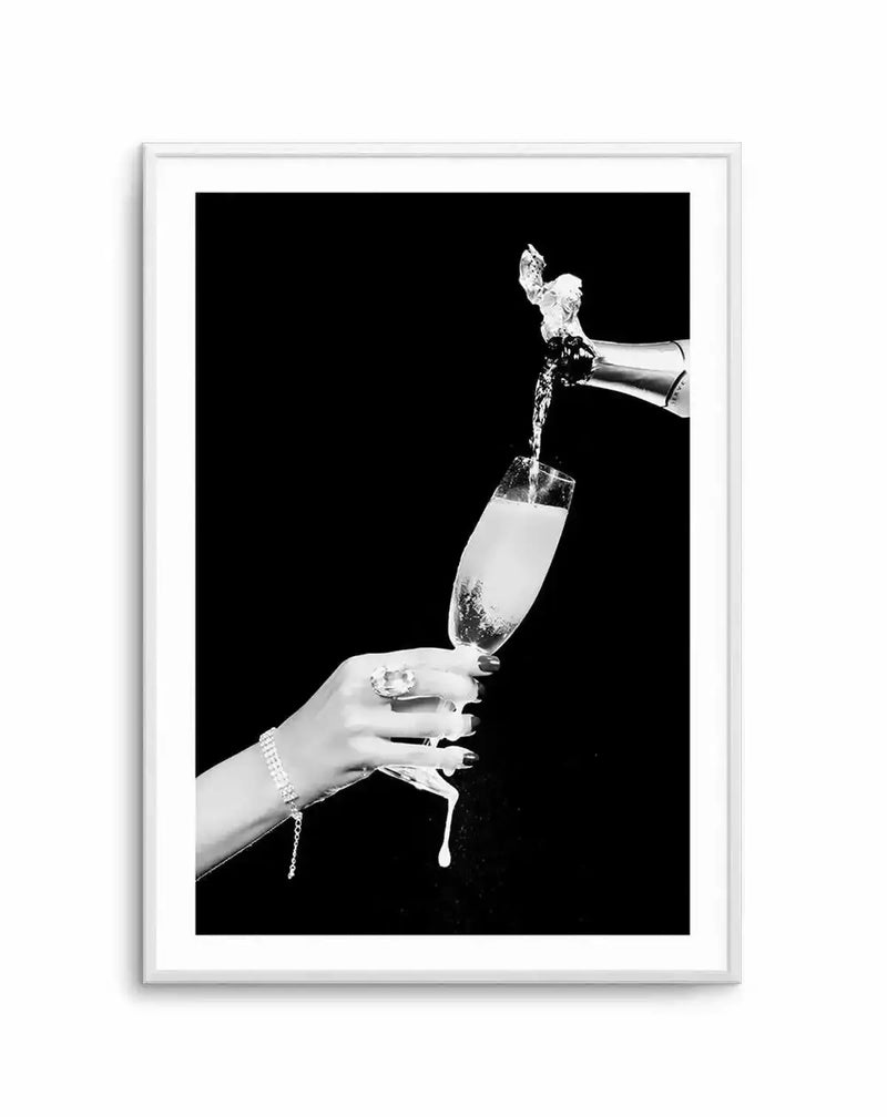 Black and white photograph showing a elegantly manicured hand holding a champagne flute while champagne is poured from a bottle above it. The hand wears a pearl bracelet and a decorative ring with a large stone. Champagne splashes dynamically into the glass against a solid black background. The artwork is printed and displayed in a silver-toned frame with a white border.
