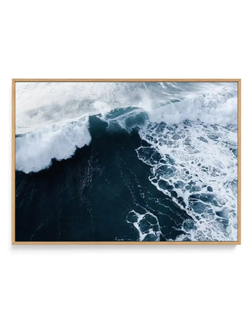 Aerial photograph of dramatic ocean waves with white foam crashing onto dark teal water, printed on canvas and framed in natural wood. The composition captures multiple wave formations from above, with churning white surf contrasting sharply against deep blue-green water. The wood frame is light tan with a clean finish, displaying the photograph in a horizontal landscape orientation.