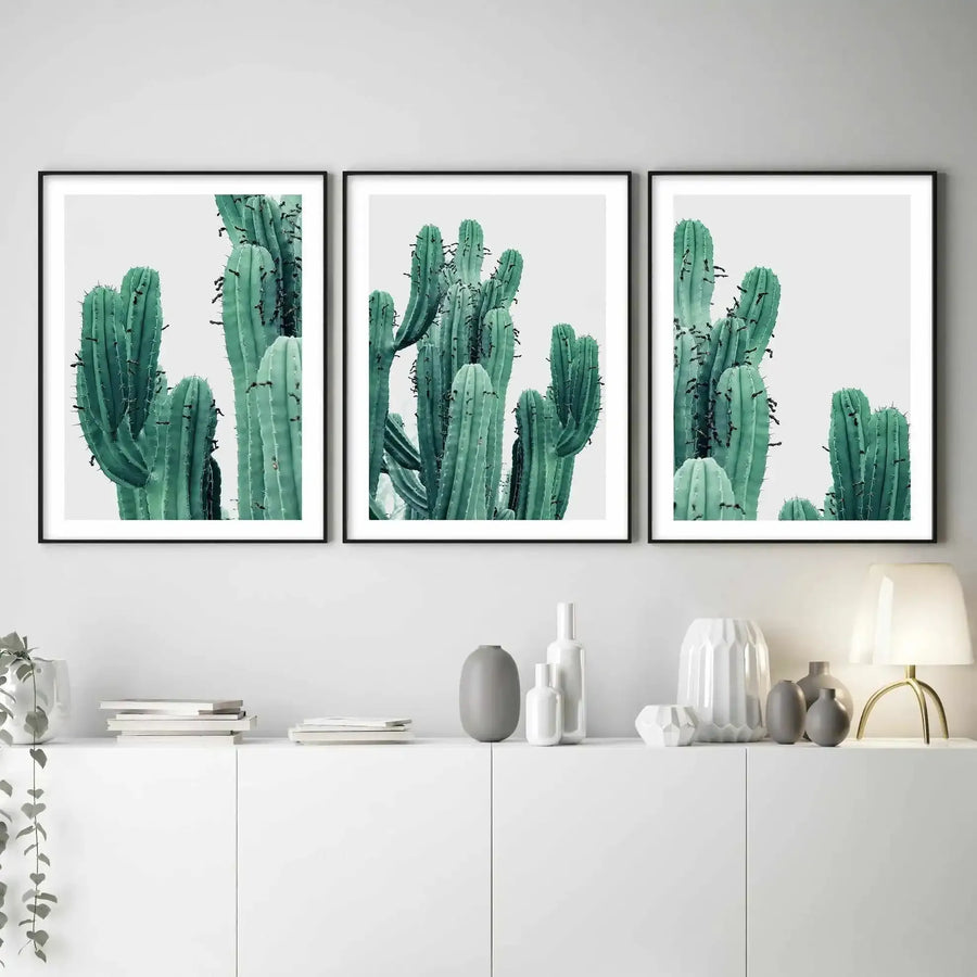 Lifestyle shot of three framed art prints displayed above a white console table in a minimalist room. Each print features a close-up photograph of tall, ribbed saguaro cacti in a muted teal-green against a stark white background, with black frames and white borders. The console table is adorned with white and grey ceramic vases, a stack of books, and a gold-toned lamp with a white shade.