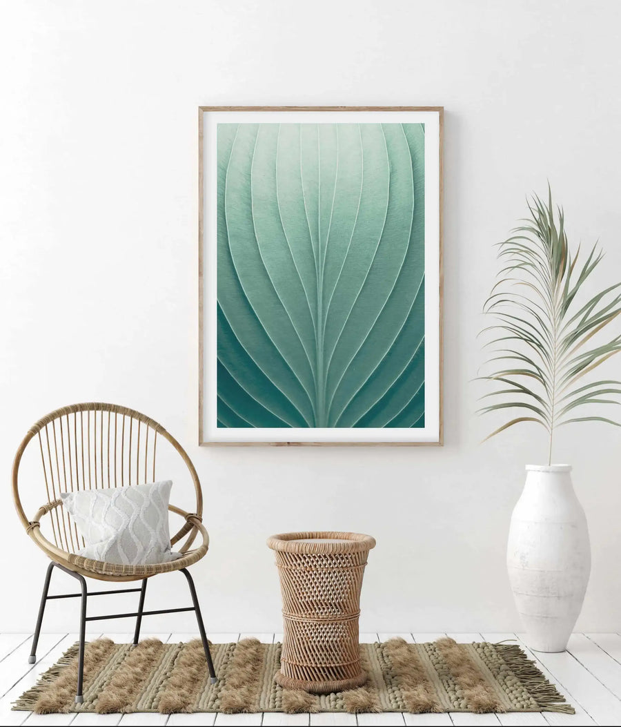 Large framed art print displayed on a white wall in a bright, minimalist living room. The print features a close-up of a green leaf with prominent veins, showcasing a gradient from light to dark teal. The print is in a natural wood frame with a white border. The room includes a round rattan chair with a white patterned cushion, a woven rattan basket, a textured beige rug, and a white vase with a dried palm frond.