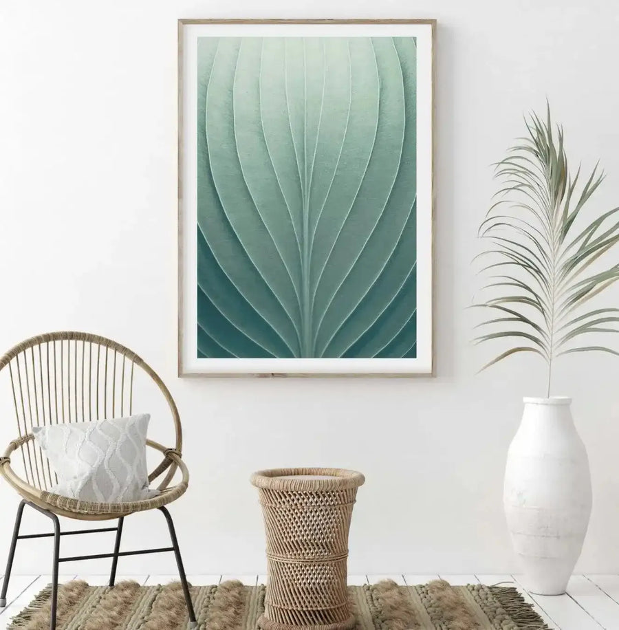Lifestyle shot featuring a framed art print displayed on a white wall in a bright, minimalist room. The print, titled "Green Leaves III," depicts a close-up, abstract view of a leaf's intricate vein structure in varying shades of teal and sage green, with a natural wood frame and white border. Below the print, a round wicker chair with a white textured cushion sits on a woven rug, next to a cylindrical wicker side table. To the right, a tall white vase holds a dried palm frond.