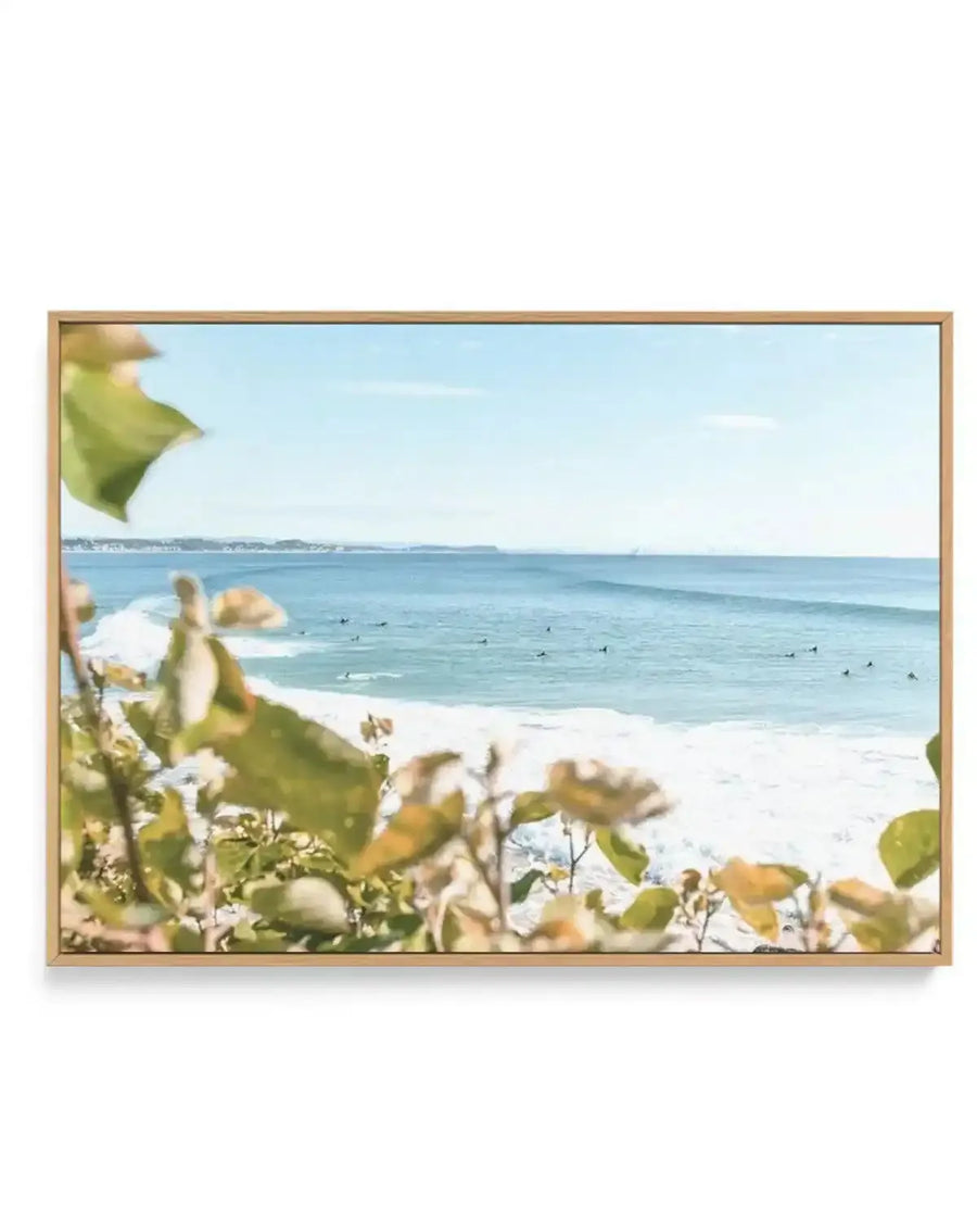 Framed canvas print of a coastal photography scene with a natural wood frame. The image captures a wide view of a light blue ocean with numerous surfers scattered across the water, waiting for waves. The foreground features blurred green and yellow-green foliage, suggesting a view from a cliff or elevated position. The sky above is a clear, pale blue with a few faint white clouds.