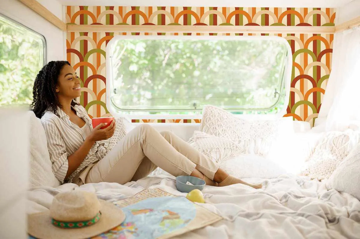 Groovy Arches Wallpaper displayed on the walls of a camper interior. The design features repeating geometric arches in burnt orange, golden yellow, olive green, and burgundy red arranged in vertical stripes against a cream background. The pattern has a retro 1970s aesthetic with bold, symmetrical half-circle motifs. The wallpaper is shown framing a large rounded window with a white border, creating a warm, cohesive look in the compact living space furnished with cream bedding and throw pillows.