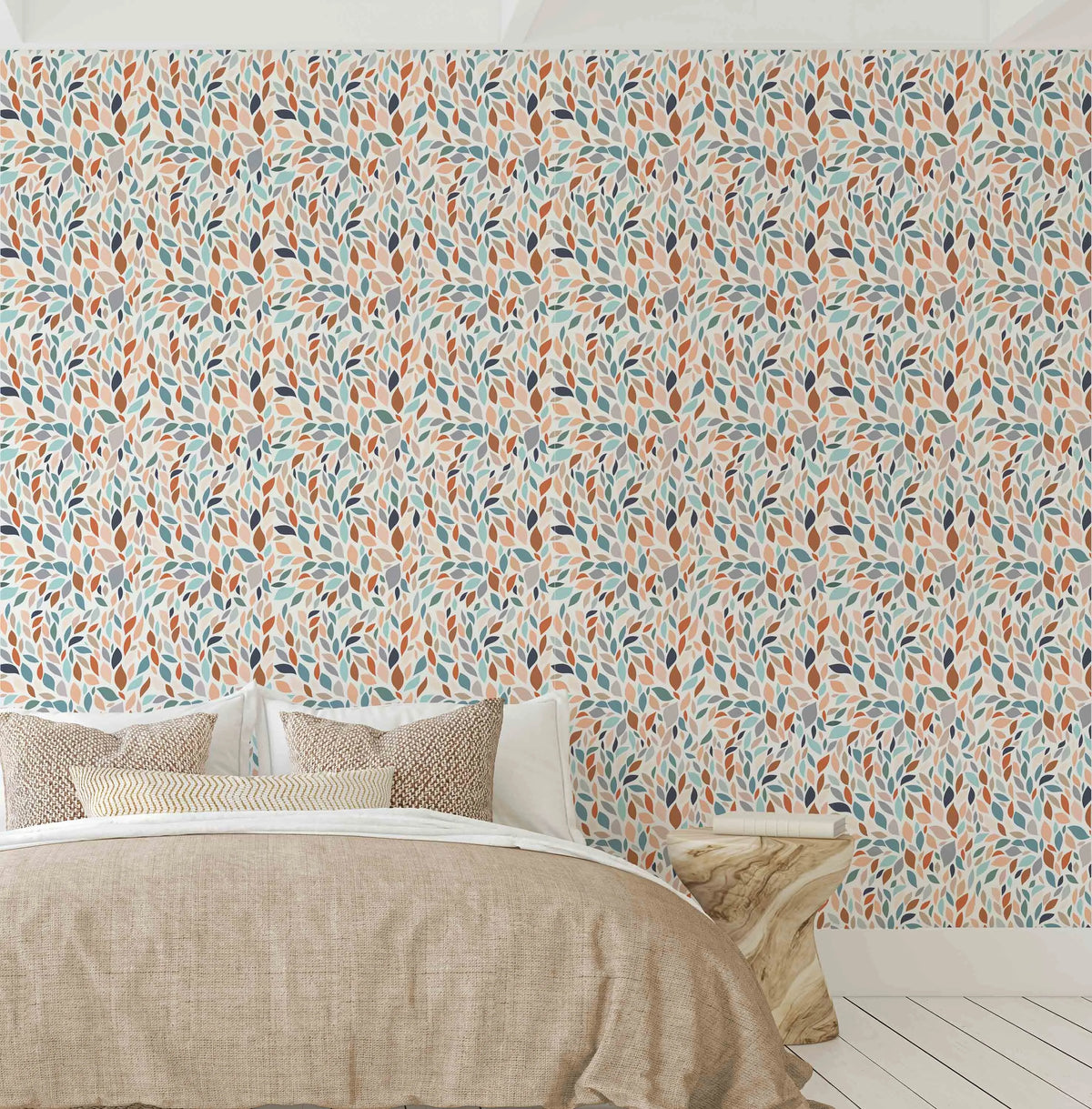 Groovy Leaves Wallpaper displayed on a bedroom wall above a bed with beige linen bedding and patterned throw pillows. The wallpaper features an all-over repeating pattern of hand-painted leaf shapes in teal, rust orange, cream, and dark navy blue on a light neutral background. The leaves are arranged in organic, flowing clusters creating a contemporary botanical design with a watercolor illustration style. The pattern covers the entire visible wall surface in a seamless, rhythmic repeat.