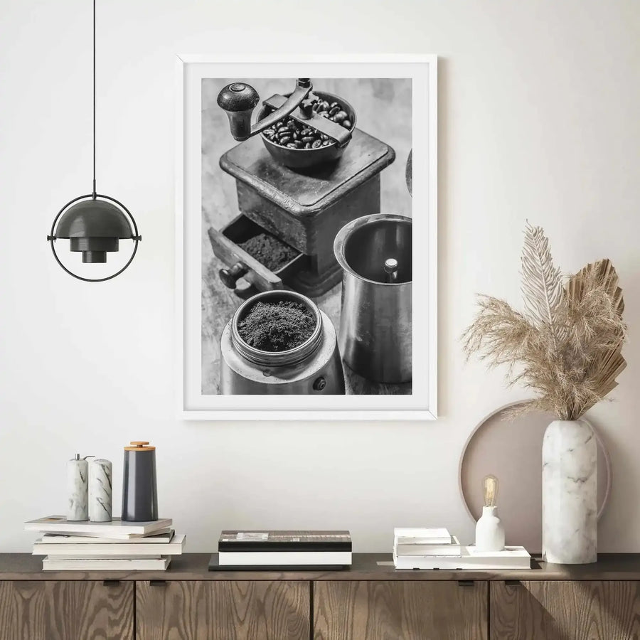 Black and white vintage photograph in white frame with white border, depicting a manual coffee grinder on a wooden base with ground coffee in a glass jar below, whole coffee beans in the grinder's hopper, and a metal container to the right. The print is displayed above a wood console table styled with stacked books, a pendant lamp, dried pampas grass in a marble vase, and decorative objects. The photograph has a grainy, documentary quality capturing classic coffee-making equipment and ingredients.