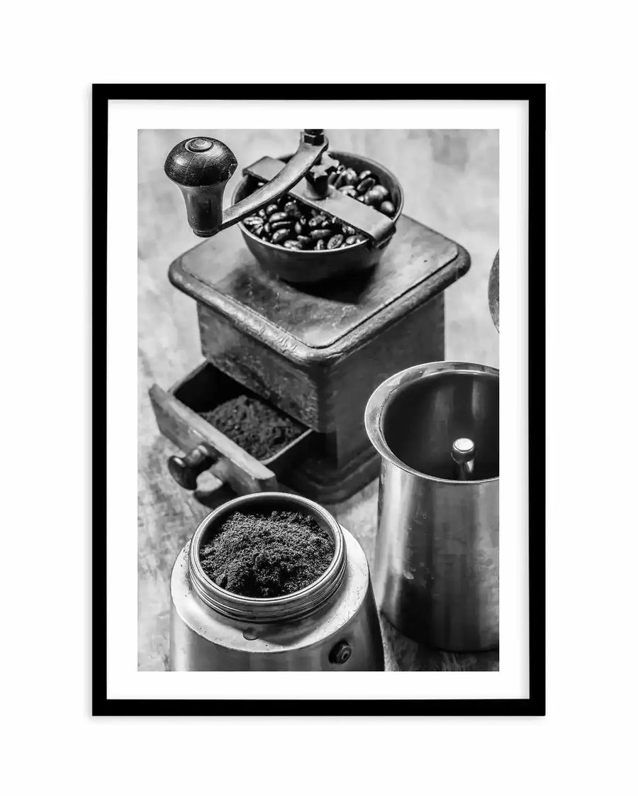 Framed black and white photograph of coffee preparation with a black frame and white border. The composition features an antique hand-crank coffee grinder with coffee beans in its hopper and ground coffee in its drawer, next to a metal moka pot base filled with ground coffee and a moka pot top.