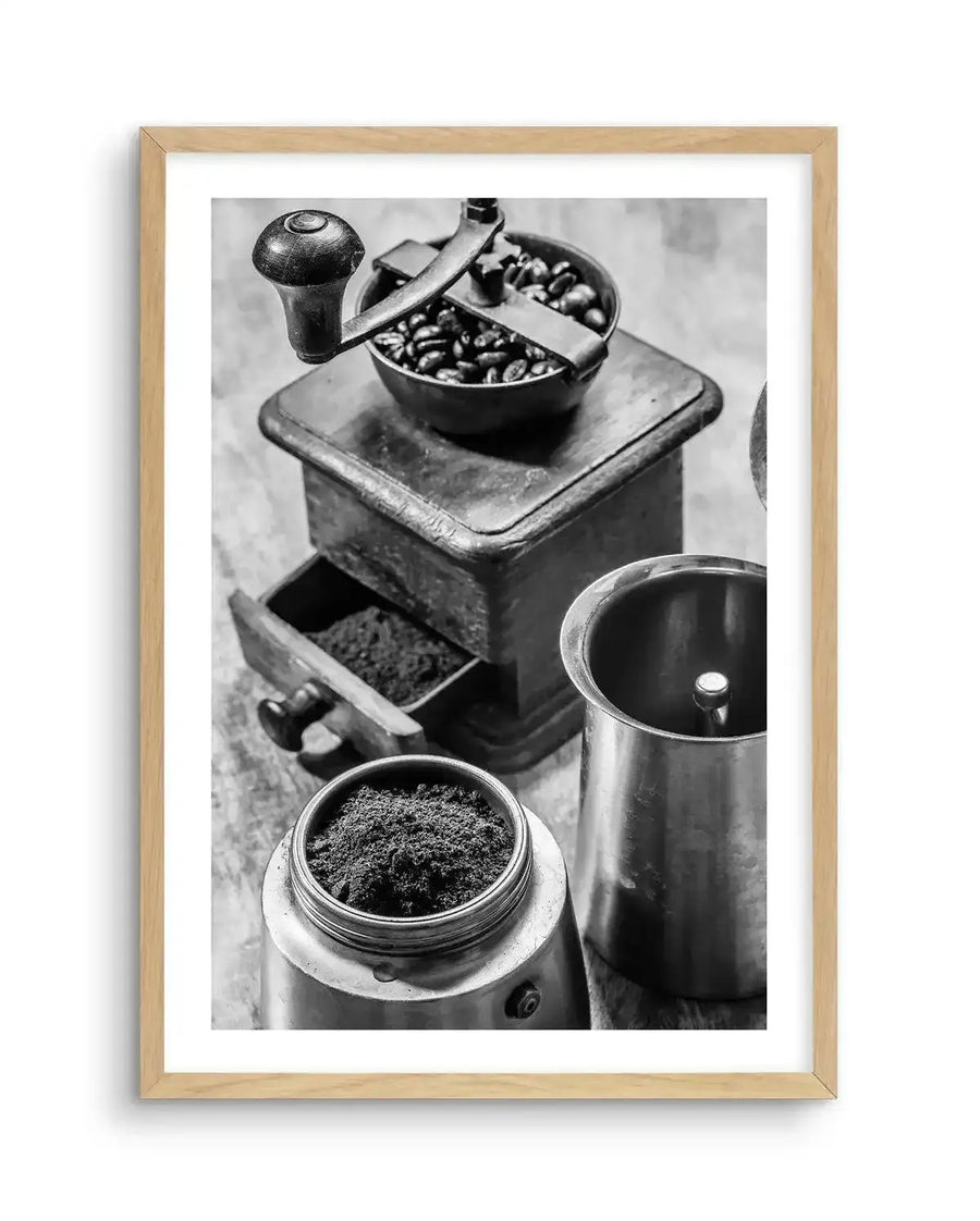 Framed black and white photograph of vintage coffee preparation tools with a natural wood frame and white border. The composition features an antique hand-crank coffee grinder filled with whole coffee beans, a drawer pulled out revealing ground coffee, and two metal moka pot components, one filled with ground coffee.