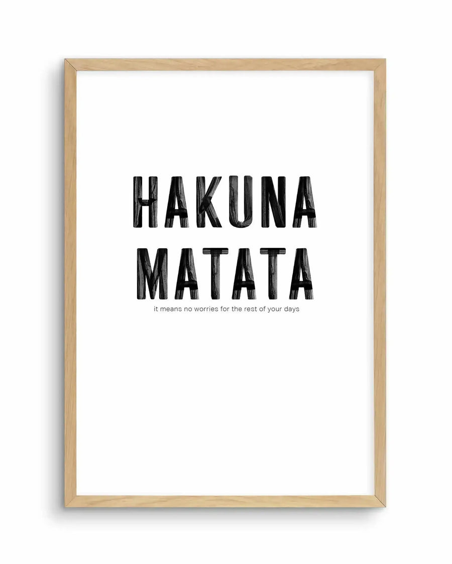 Framed art print with light wood frame displaying "HAKUNA MATATA" in large black capital letters with a sketched texture, centered on white background. Below the main text in smaller black letters reads "it means no worries for the rest of your days." Minimalist design with high contrast between bold typography and clean white space.