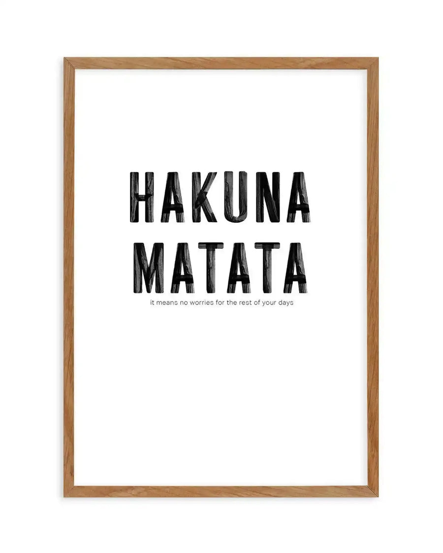 Art print featuring the words "HAKUNA MATATA" in large, distressed black capital letters, with "it means no worries for the rest of your days" in smaller black text below, displayed in a natural oak frame with white border.