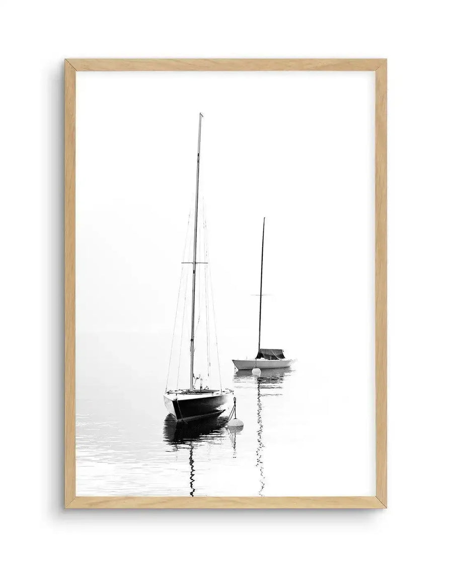 Framed black and white photograph of two sailboats moored on calm water with subtle reflections, displayed in a natural oak frame with white border. The composition features a larger sailboat in the foreground and a smaller one behind, both with tall masts, against a bright, minimalist background.