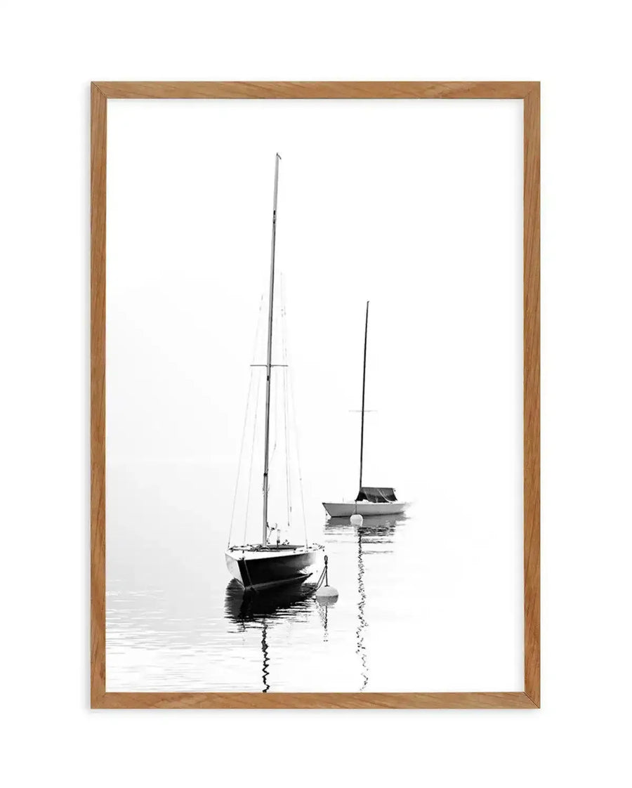 Framed black and white photograph of two sailboats anchored on calm water, displayed in a natural oak frame with white border. The composition features the larger sailboat in the foreground with its mast reaching towards the top of the frame, and a smaller sailboat in the background. Both boats are reflected in the still water, creating a serene and minimalist scene.