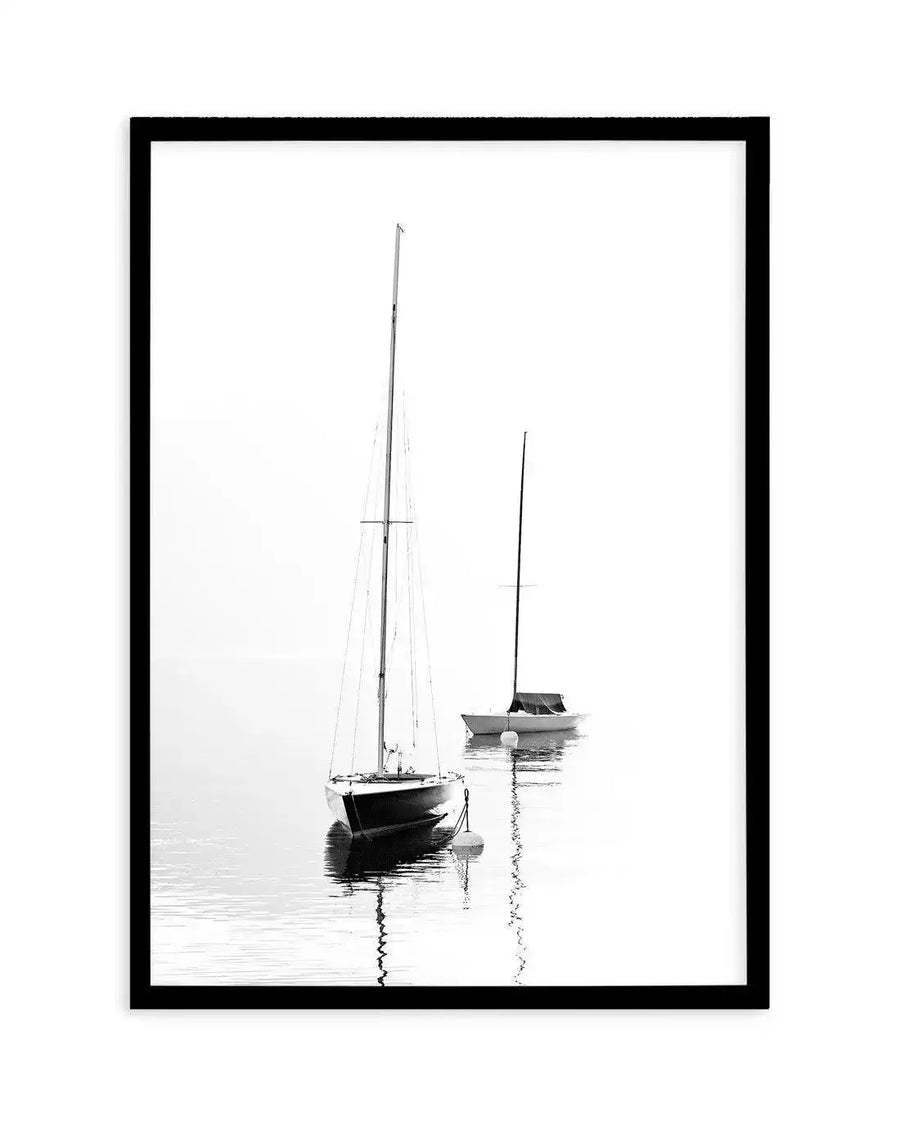Framed black and white photograph of two sailboats moored on calm water with a soft, hazy background, displayed in a black frame with white border. The foreground boat is larger and darker, reflecting in the water, while the smaller boat behind it appears lighter and more distant.