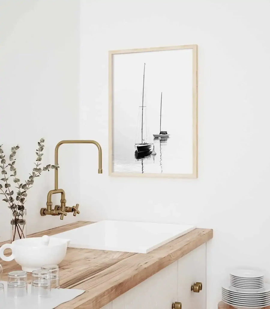 Lifestyle shot of a minimalist kitchen featuring a framed black and white photograph of two sailboats on calm water, displayed above a white sink. The print has a natural wood frame with a white border. The kitchen has white walls, a light wood countertop, a brass faucet, and a vase with dried eucalyptus branches.