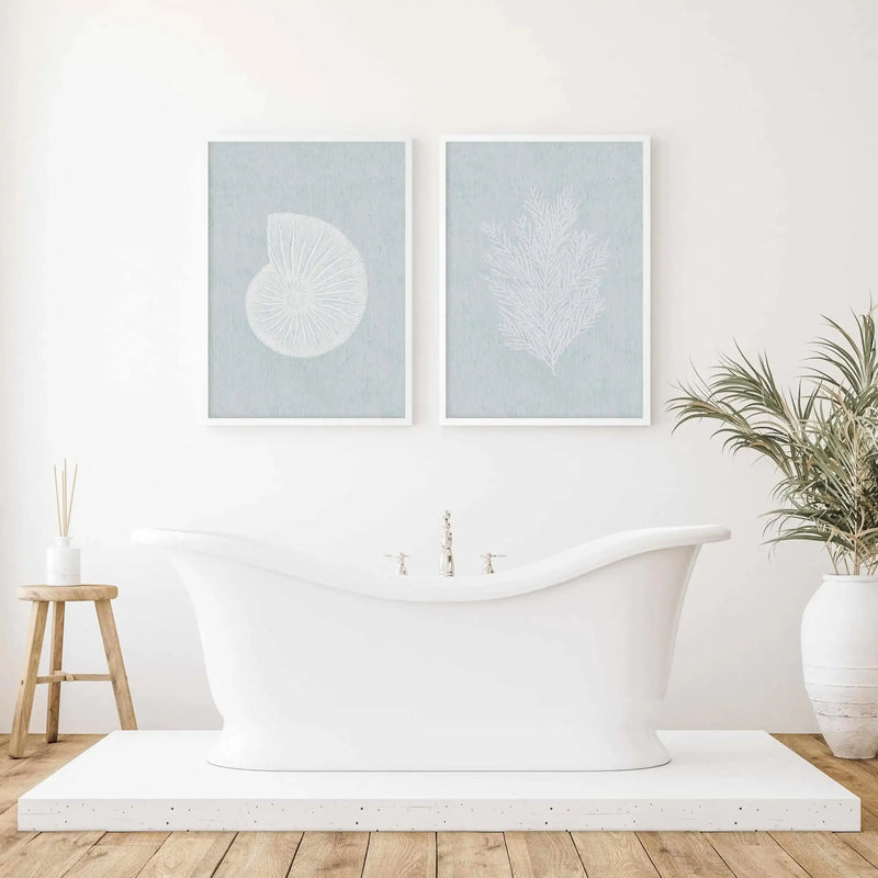 Two framed art prints displayed above a white freestanding bathtub in a spa-like bathroom setting. Left print features a white circular shell or fan design on soft sage green background. Right print shows white feathery palm fronds on matching sage green background. Both prints have white frames with white borders. Wooden stool with decorative diffuser and potted palm plant flank the bathtub. Light wood flooring and white walls complete the serene coastal aesthetic.