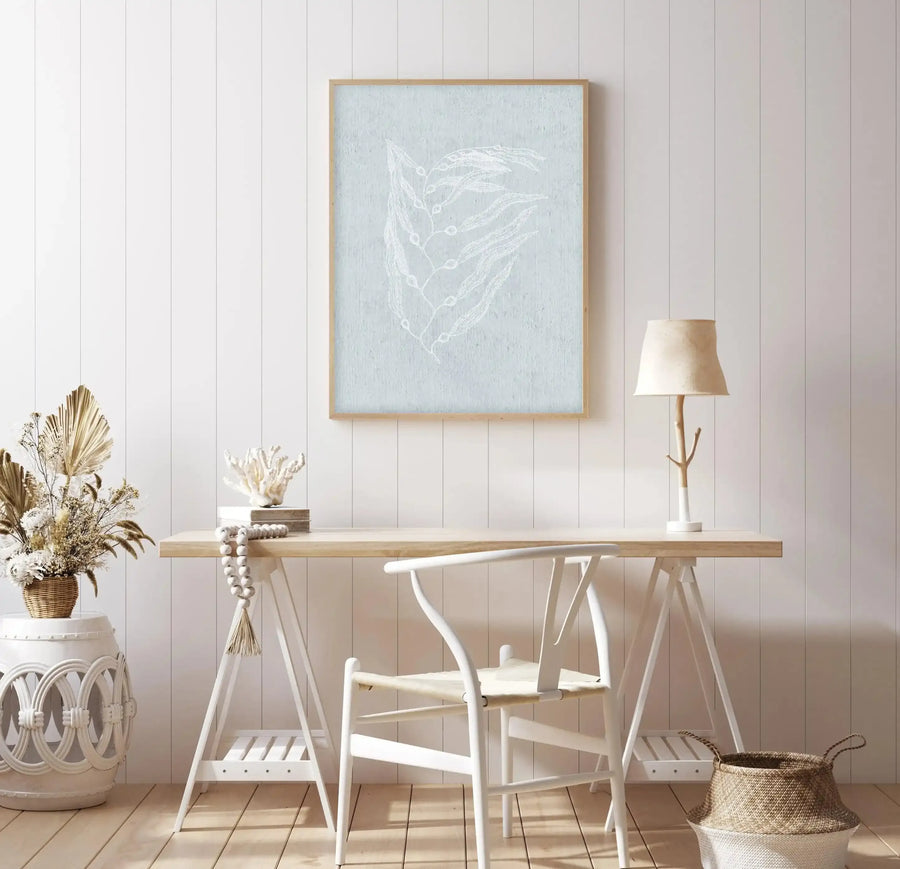 Framed art print displaying a delicate white botanical leaf illustration on a soft sage green background, presented in a natural wood frame. The print hangs on a white shiplap wall above a wooden desk with white chairs in a bright, minimalist Hamptons-style interior featuring dried pampas grass, a table lamp, and woven baskets.