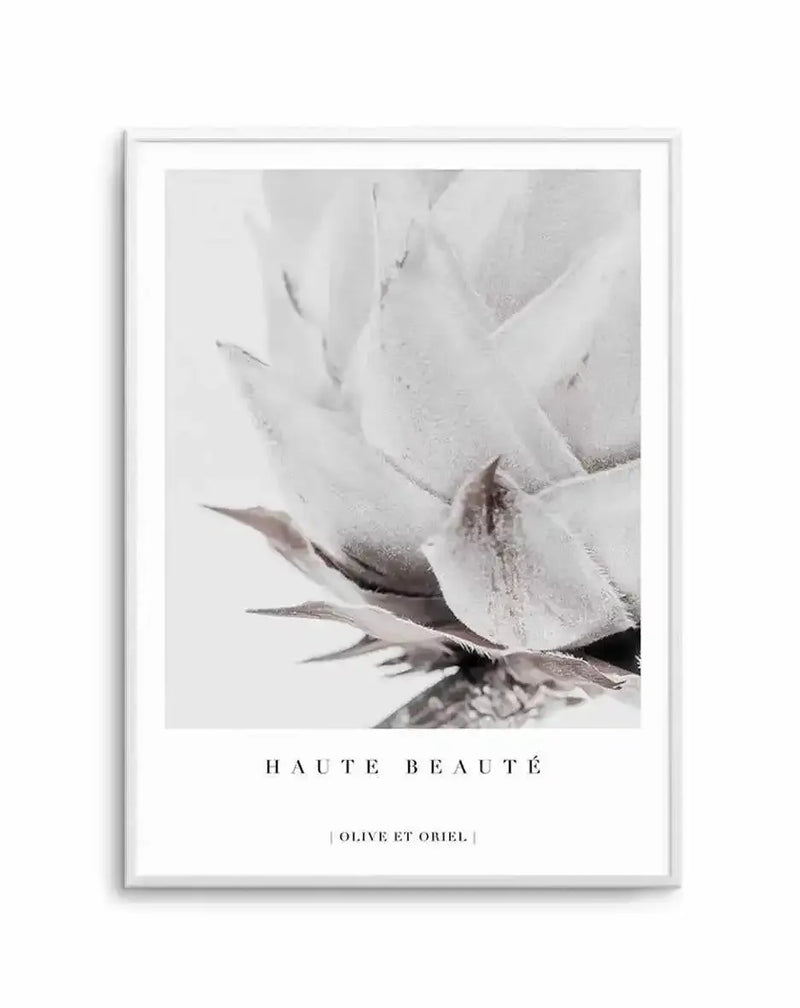 Framed art print featuring a close-up black and white photograph of a king protea flower. The image shows delicate white petals with soft gray and taupe tones, capturing fine textural details and the flower's intricate center. The photograph has a minimalist, ethereal quality with subtle shadows and gentle gradations. Text below reads "HAUTE BEAUTÉ" and a small credit line. The artwork is mounted in a white frame with white border against a neutral background.