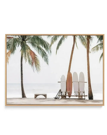 Framed canvas art print featuring a beach scene with four surfboards in various shades of white, cream, and rust orange leaning against a metal rack between two tall palm trees on sand. A wooden bench sits to the left. The ocean and overcast sky are visible in the background. The photograph has a muted, natural color palette with soft lighting. The canvas is mounted in a light natural wood frame with white border.