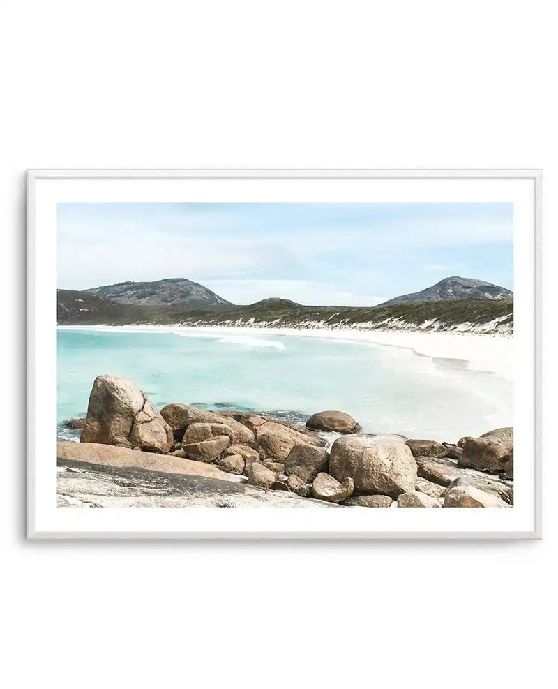 Framed photograph of a serene coastal beach scene with a white frame and white border. The image features clear turquoise water gently meeting a wide expanse of white sand, with a rocky foreground. In the distance, rolling hills covered in sparse green vegetation rise against a soft blue sky with light clouds.