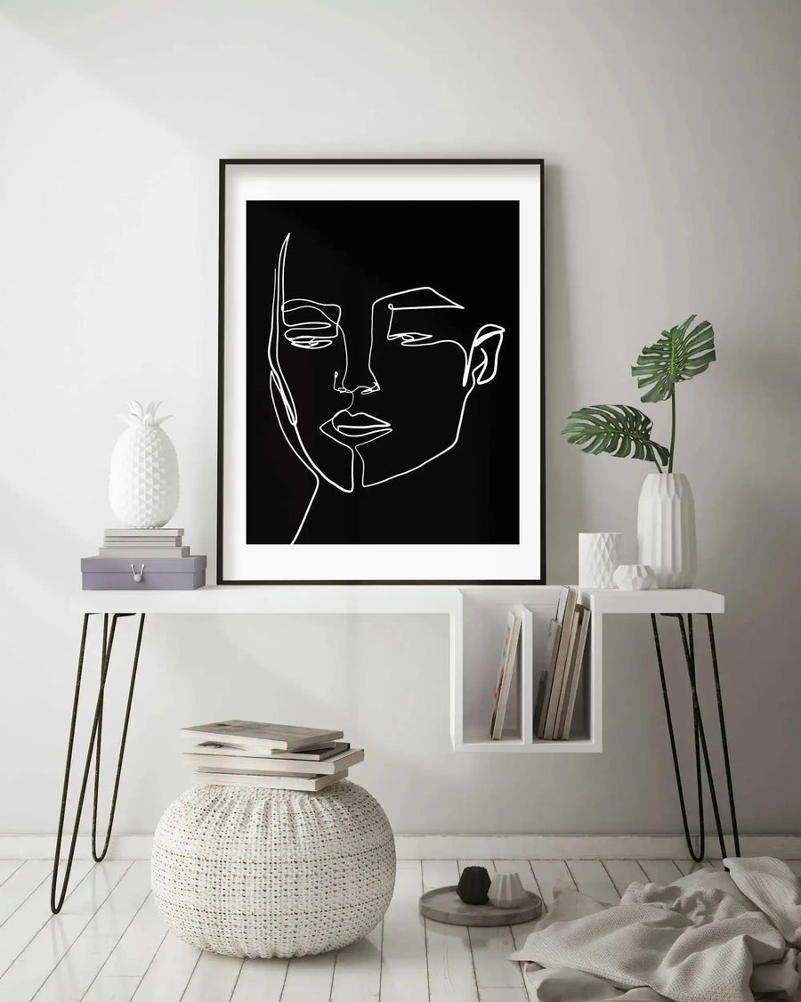 Lifestyle shot of a minimalist room featuring a framed art print of a woman's face in a continuous line drawing style, displayed above a white console table. The print is black and white with a black frame and white border. The room has white walls and light wood flooring, with a white pineapple sculpture, grey books, and a monstera plant in a white vase on the table. A white knitted pouf with books sits on the floor next to the table.