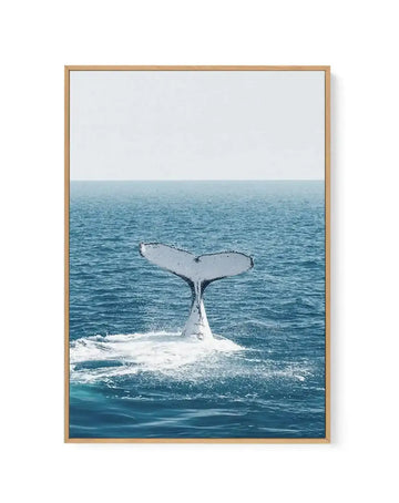 Framed canvas art print featuring a photograph of a whale's tail breaching in ocean waves. The composition shows a white and dark grey whale tail emerging from turquoise and teal blue water with white foam, set against a soft grey-blue horizon and pale sky. The canvas is mounted in a natural wood frame with a white border, creating a vertical rectangular format suitable for wall display.
