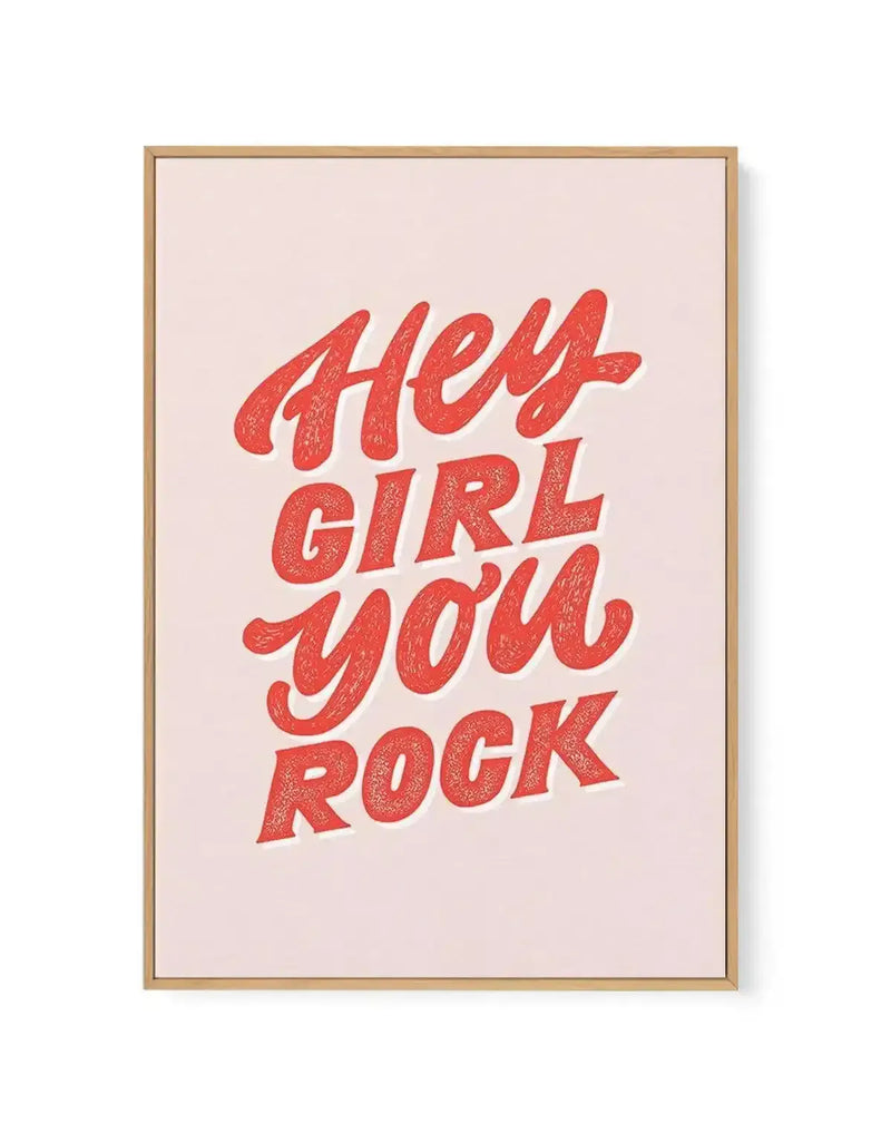 Framed canvas art print with a natural wood frame displaying the text "HEY GIRL YOU ROCK" in bold, textured red lettering with a distressed, speckled finish against a soft beige background. The design uses a playful, retro-inspired script font with organic, curved letterforms. White border surrounds the artwork inside the frame.