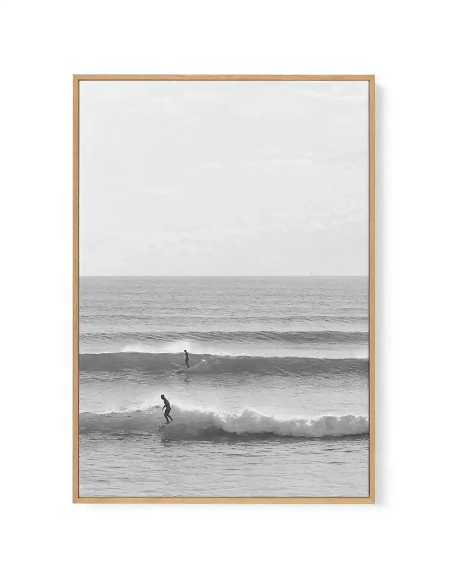 Framed black and white photograph with natural wood frame showing two surfers in wetsuits riding waves on a misty, overcast day. The composition is vertical with calm gray sky in the upper half and layered ocean waves in the lower half. The photograph has a serene, minimalist quality with soft tonal gradations. White border surrounds the image within the light wood frame.