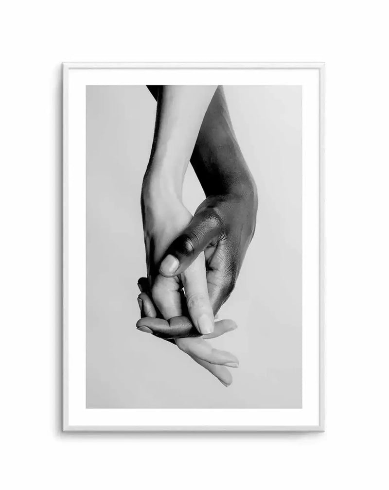 Framed black and white photograph of two hands, one light-skinned and one dark-skinned, holding each other gently. The light-skinned hand is on the left, and the dark-skinned hand is on the right, with fingers intertwined in a tender embrace, displayed in a white frame with white border.